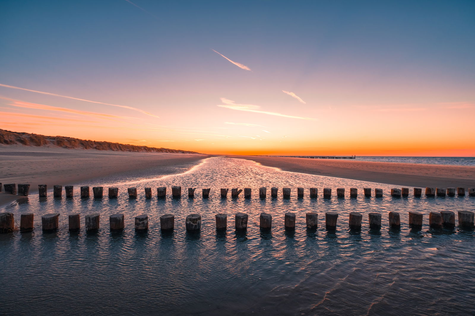 Sunset on a beach with two parallel rows of wooden groynes reflected in the calm, shallow water. A sandbank stretches towards the orange horizon, with dunes to the left under a sky that fades from deep blue to warm orange.